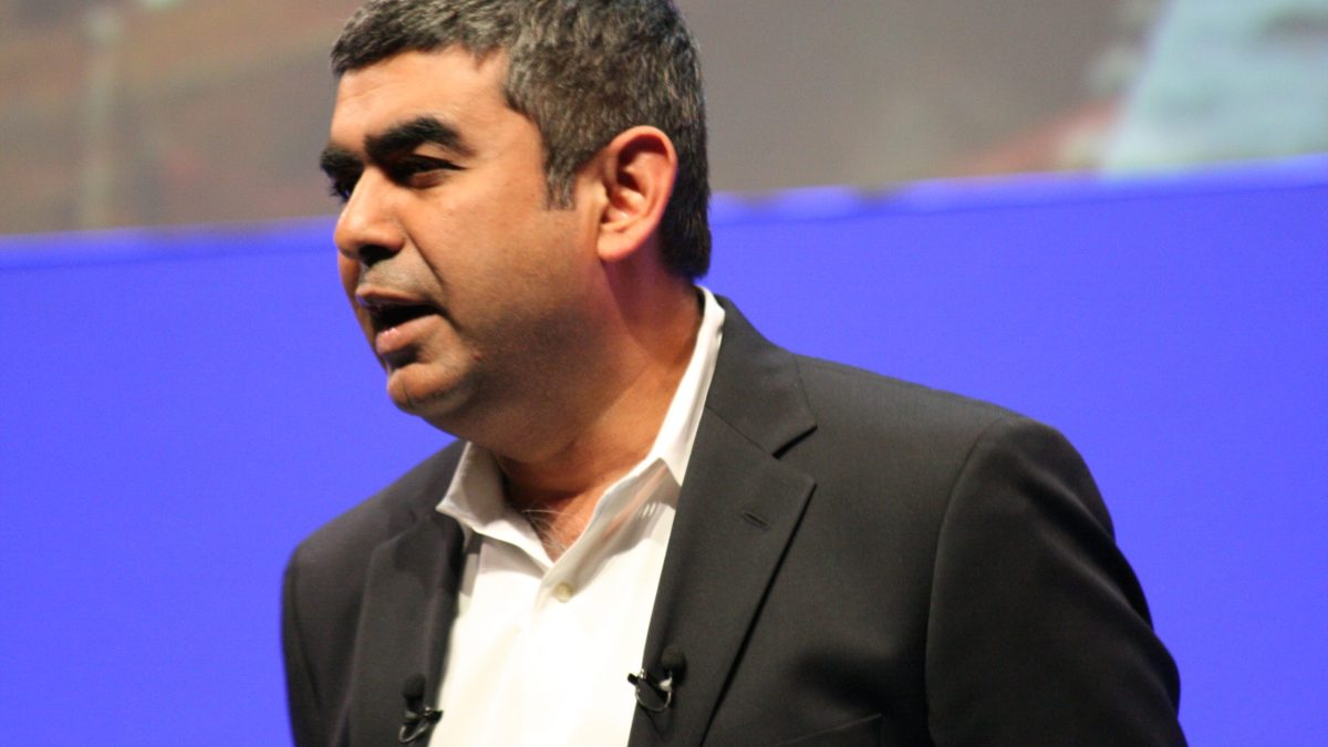 Vishal Sikka in a dark jacket speaking on stage against a blue background