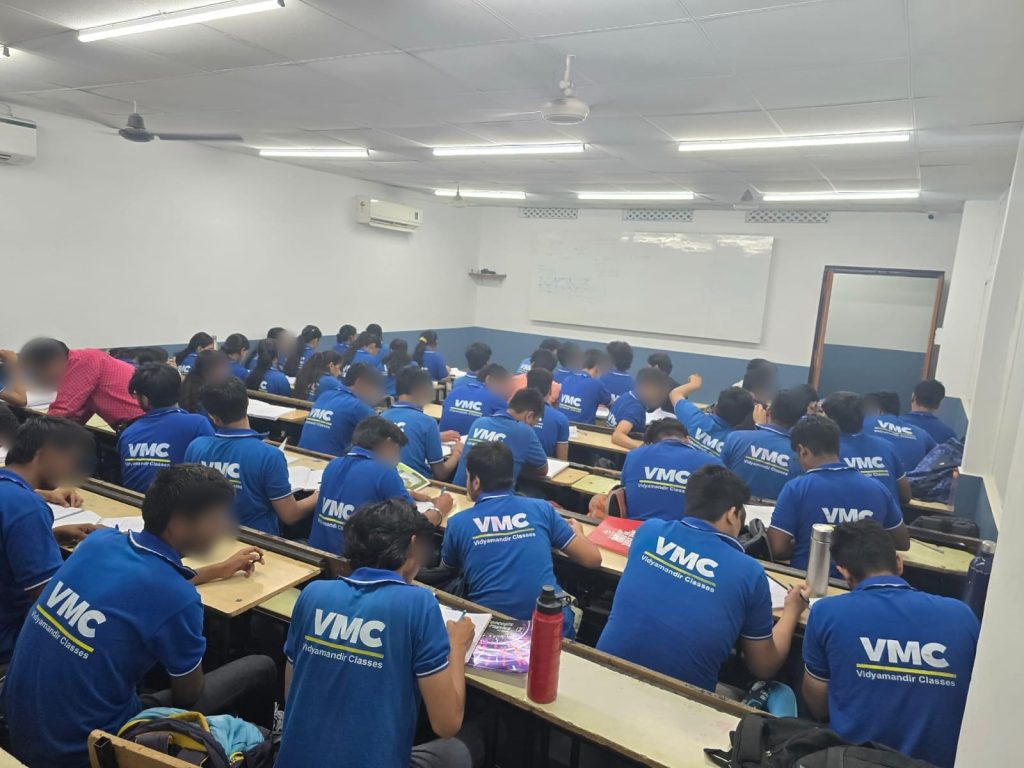 Students in blue Vidyamandir Classes uniforms seated in a classroom, seen from behind