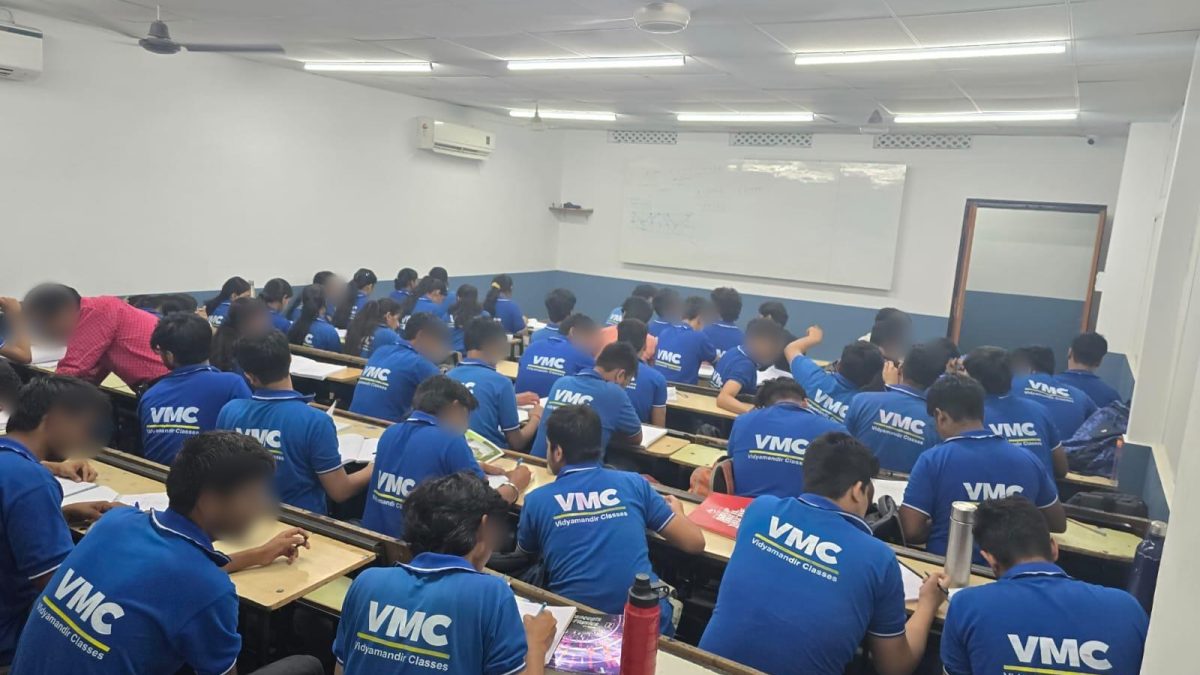 Students in blue Vidyamandir Classes uniforms seated in a classroom, seen from behind