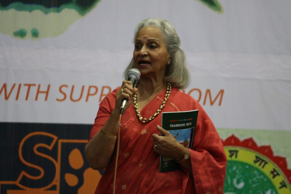 Waheeda Rehman with white hair, in a red patterned sari, speaking into a microphone