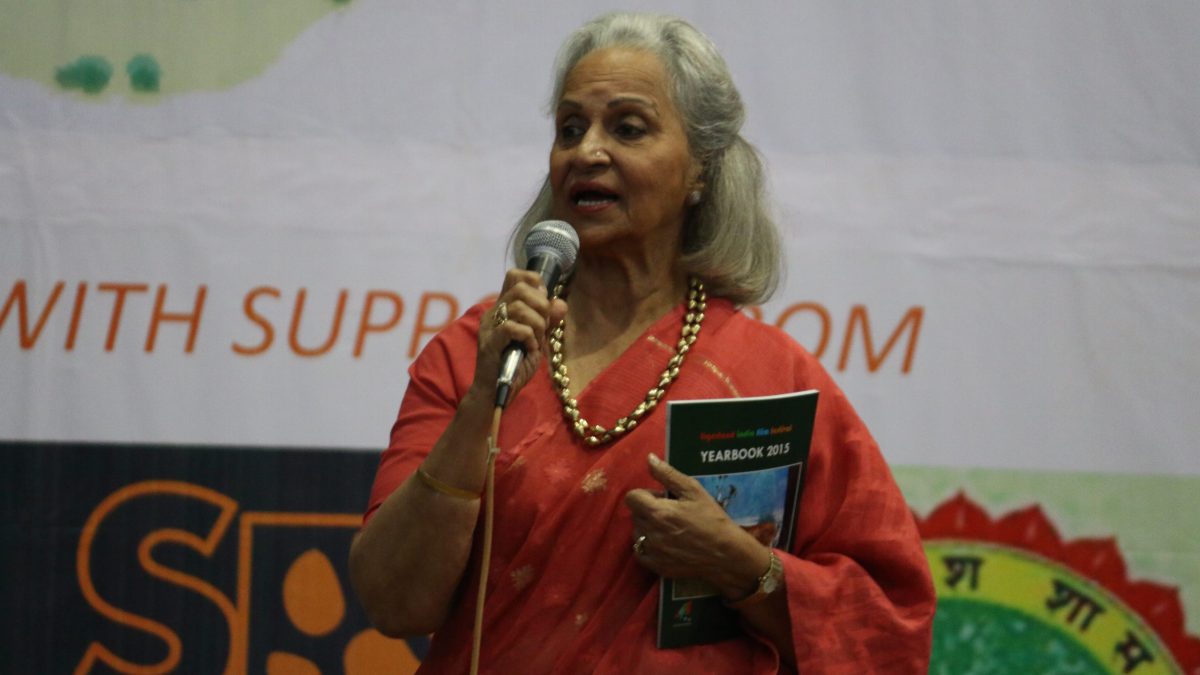 Waheeda Rehman with white hair, in a red patterned sari, speaking into a microphone