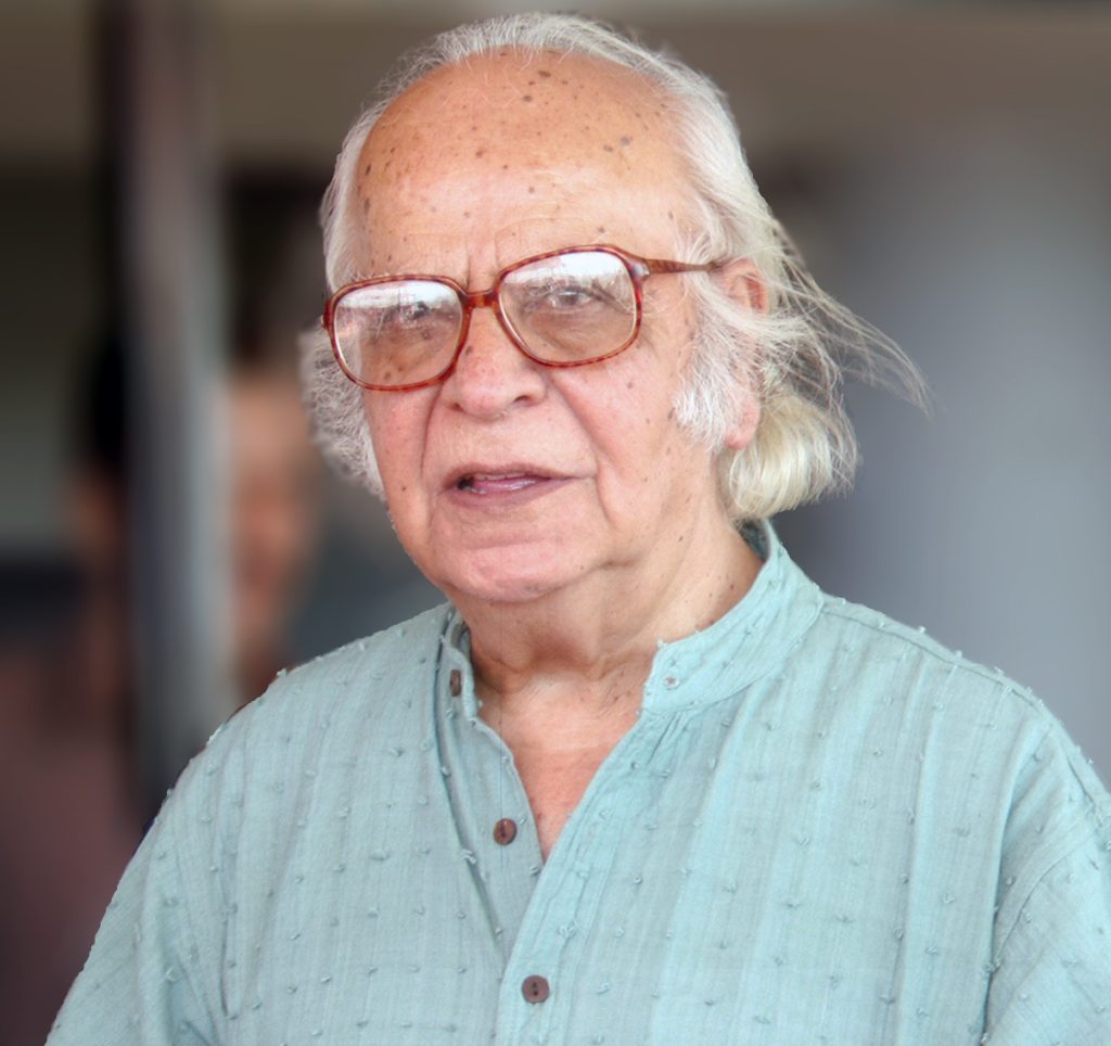 Yash Pal with white hair and glasses, in a pale kurta, speaking