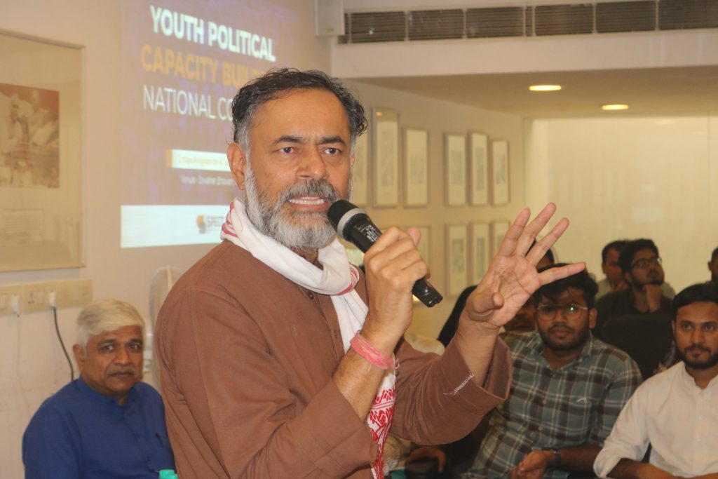 Yogendra Yadav with a grey beard speaking into a microphone and gesturing to a seated audience