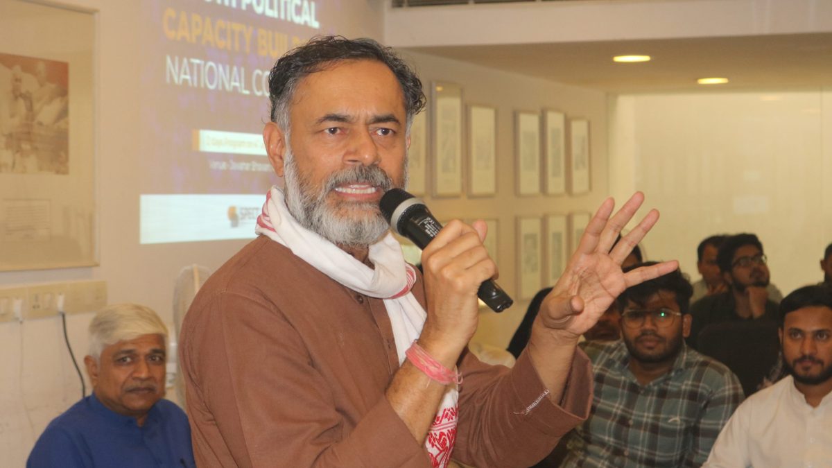 Yogendra Yadav with a grey beard speaking into a microphone and gesturing to a seated audience