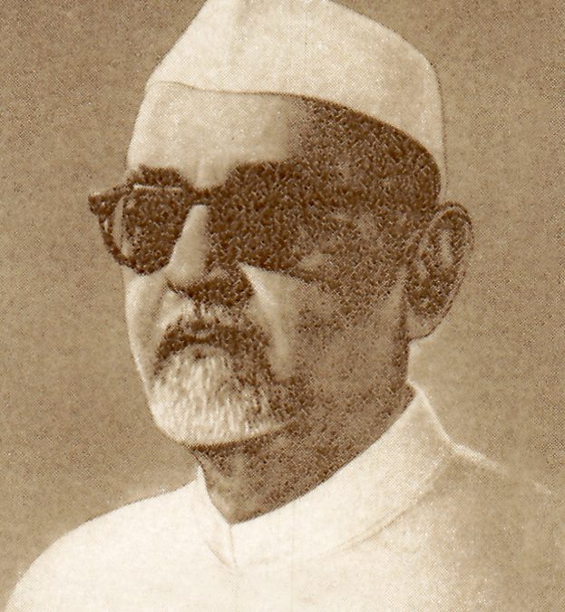 A portrait of Zakir Husain in a white cap and dark glasses, from a postage stamp