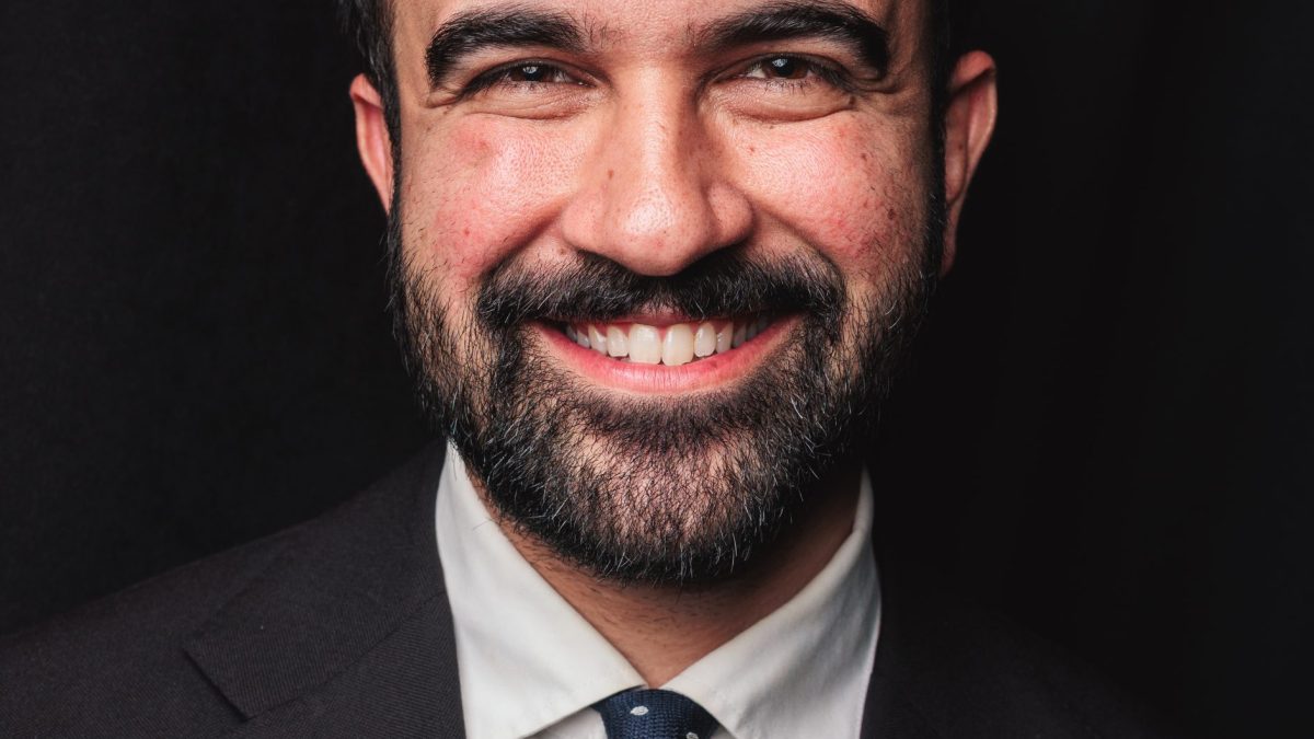 Zohran Mamdani with a beard, in a suit, smiling