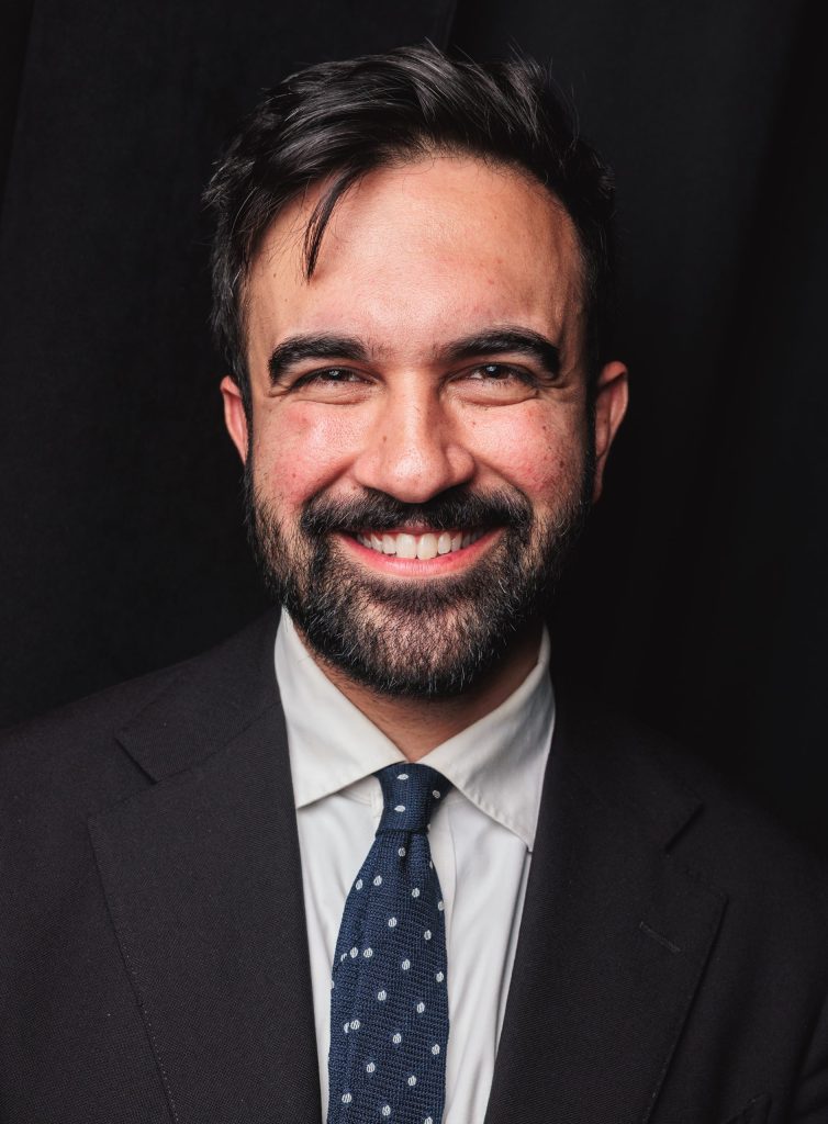 Zohran Mamdani with a beard, in a suit, smiling